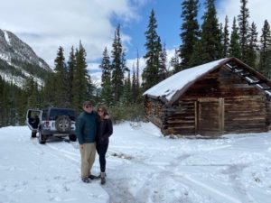 A couple stands in snow outside a mountain cabin