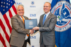 Two men shake hands while holding an award in front of a FEMA flag and US flag