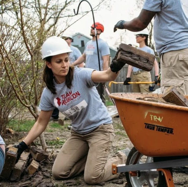 Lisa Smith volunteering with Team Rubicon - she is putting logs and other debris in a wheelbarrow while wearing a hard hat. 